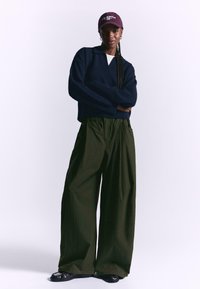 Navy knitted sweater with v-neck, paired with loose olive green wide-leg pants. Model wears a burgundy cap and black shoes.