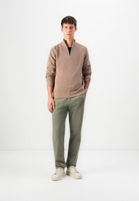 Junger Mann steht mit den Händen in den Taschen, trägt einen beigen Strickpullover, olivgrüne Hosen und hellbeige Sneakers vor einem weißen Hintergrund.