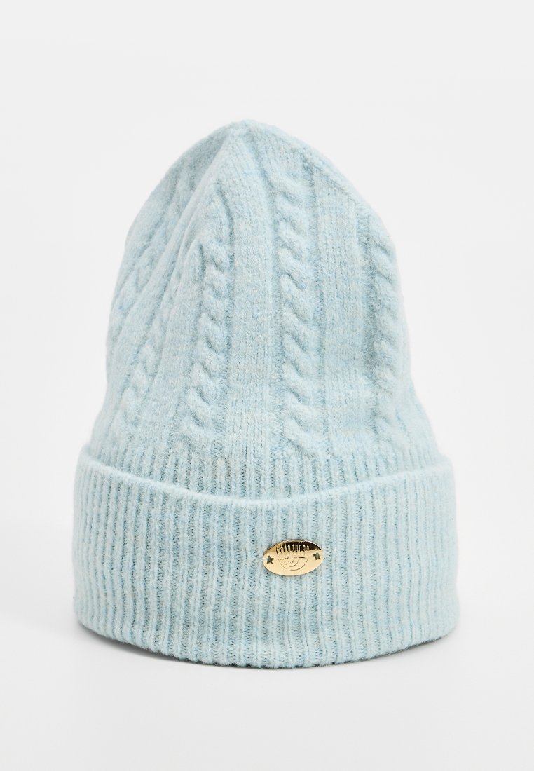Helle blaue gestrickte Wintermütze mit Zopfmuster und Rippenbündchen, versehen mit einem kleinen ovalen goldfarbenen Metallemblem an der Vorderseite.