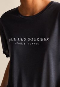 Dunkelgraues T-Shirt mit weißem Text "RUE DES SOURIRES - PARIS, FRANCE" in einer einfachen Serifenschrift; glatte Textur.