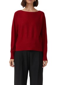 Roter Strickpullover mit weiter Boot-Ausschnitt, gerippter Textur am Saum und an den Manschetten sowie einem leicht lockeren Schnitt, kombiniert mit schwarzen Hosen.