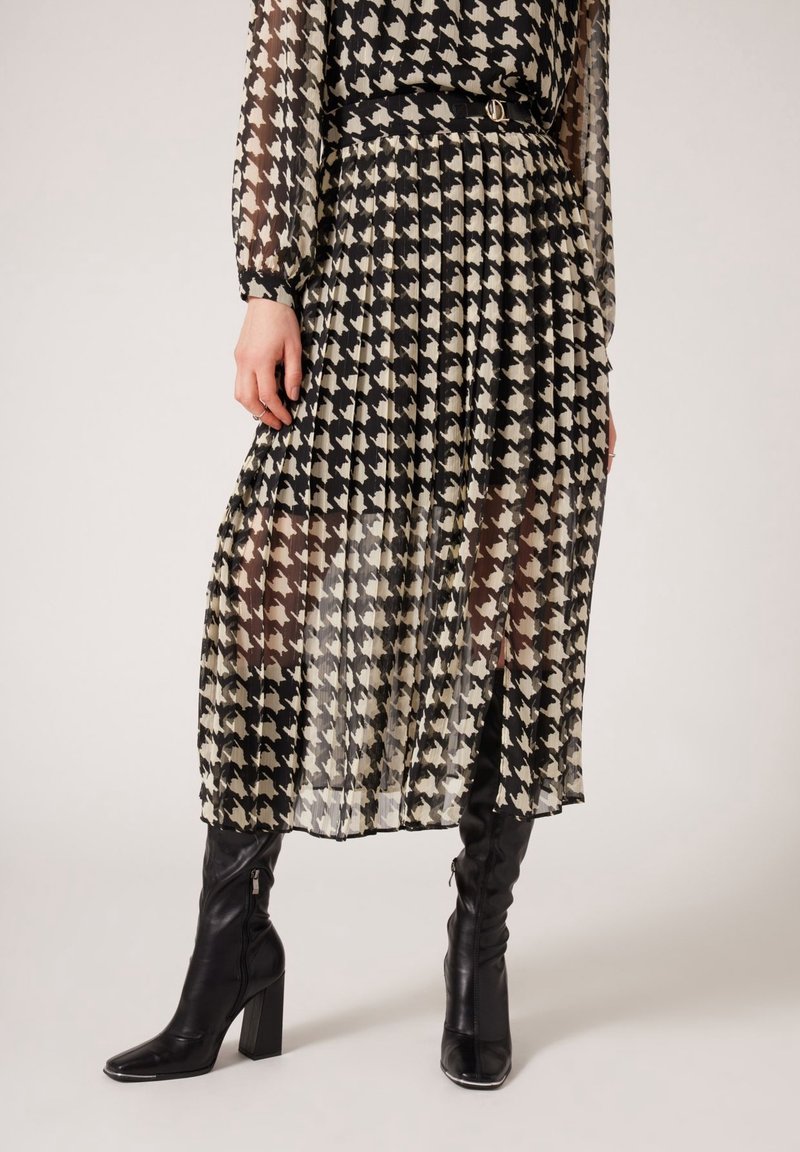 Femme portant une jupe plissée pied-de-poule noire et blanche en tissu transparent et des bottes noires hautes à talons.