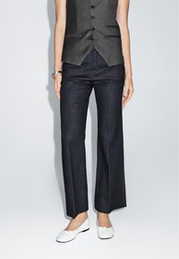 Dark denim flared pants with a smooth texture, paired with a fitted gray vest. The footwear is white, simple ballet flats.