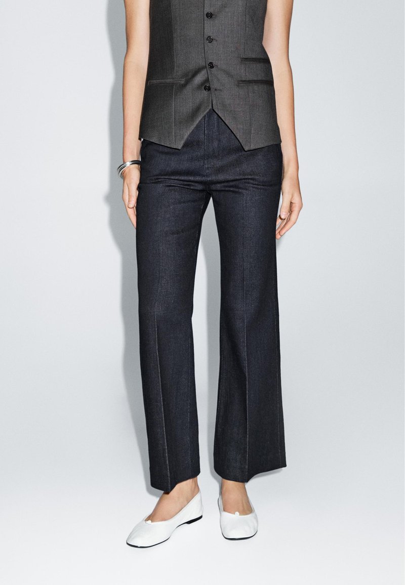 Dark denim flared pants with a smooth texture, paired with a fitted gray vest. The footwear is white, simple ballet flats.