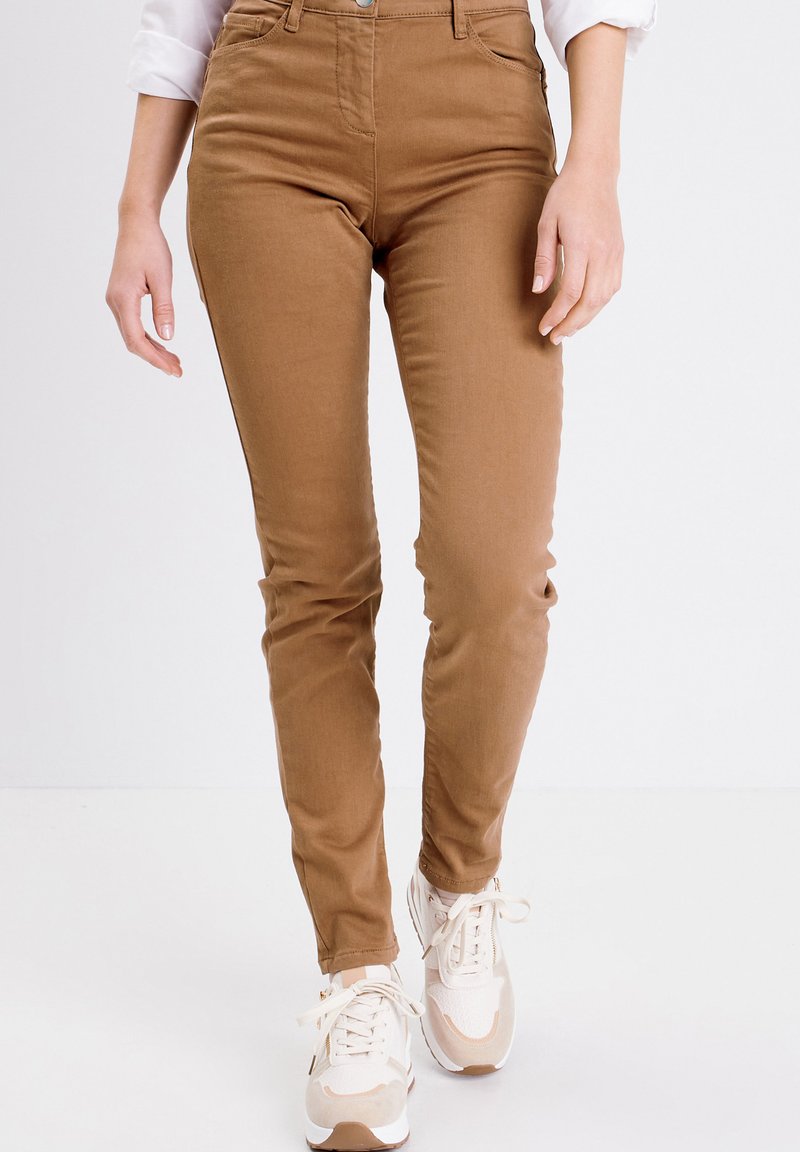 Pantalons taille haute brun clair en tissu doux en mélange de coton, présentant une coupe slim et des poches avant. Associés à des baskets beiges.