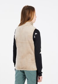 Beige Fleece-Weste mit hohem Kragen und ärmellosem Design. Über einem schwarzen Langarmshirt mit weißem geometrischen Muster getragen. Leicht strukturierter Stoff.
