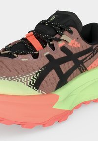 Chaussure de course marron clair et noire avec des motifs en zigzag, semelle intermédiaire vert fluo, semelle extérieure corail et lacets noirs avec des détails réfléchissants.