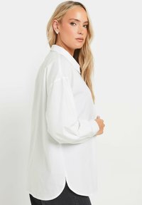 Camicia bianca con bottoni realizzata in tessuto leggero, dotata di colletto classico, maniche lunghe e orlo curvo con leggere fessure ai lati.
