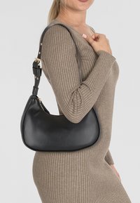Borsa a spalla in pelle nera con forma curva, hardware dorato e tracolla staccabile. Texture liscia con un sottile dettaglio di branding.