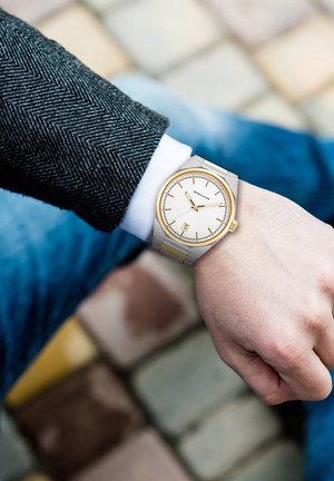 Reloj en tonos plateado y dorado con un dial texturizado de color gris claro, marcadores de hora minimalistas y función de fecha en una correa metálica, usado en una muñeca.