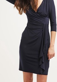 Nainen, joka on pukeutunut tummansiniseen, polvipituiseen muotovalintaan, jossa on kolmivartiset hihat ja röyhelöyksityiskohta sivussa.