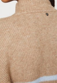 Gros plan sur un pull à col roulé beige en maille avec une petite étiquette ronde en métal près du col à l'arrière.