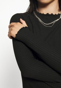 Černý žebrovaný top s dlouhými rukávy, který má vykrojený výstřih a manžety. Materiál má hladkou texturu a přiléhavý design.