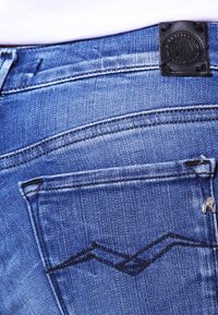 Pantalones vaqueros de denim en azul oscuro con un ligero desgaste. Presentan un parche de cuero negro en la cintura y costuras decorativas en el bolsillo trasero.