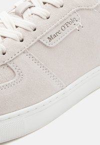 Hellgrauer Veloursledersneaker mit weißer Gummisohle, Schnürsenkeln und einem Logo-Patch mit "Marc O'Polo"-Emblem an der Seite. Detail mit glatter Textur.