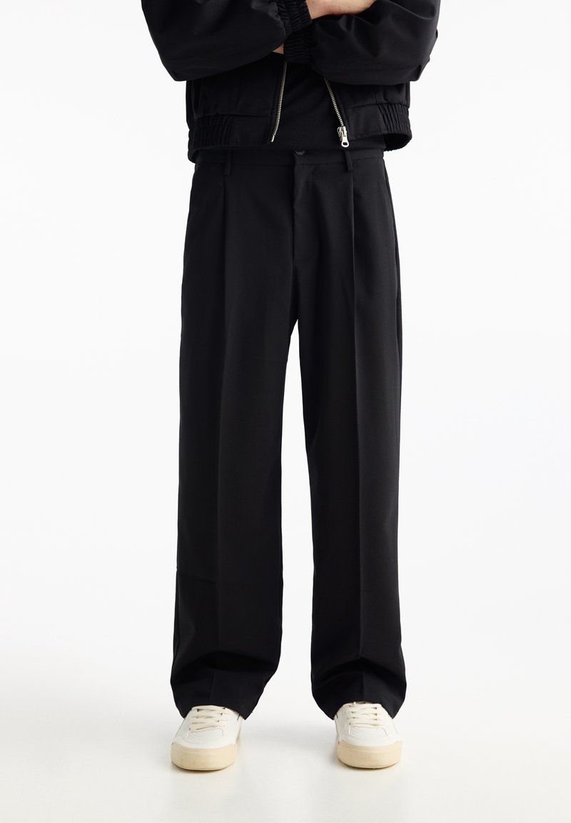 Pantalon noir à jambes larges avec une texture lisse et des plis à l'avant. Associé à des baskets blanches, avec une coupe décontractée et des détails subtils.