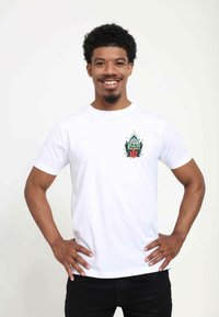 Hvid bomulds t-shirt med et farverigt grafisk design på venstre bryst, der viser en karakter blandt blomsterdetaljer. Korte ærmer, klassisk pasform.