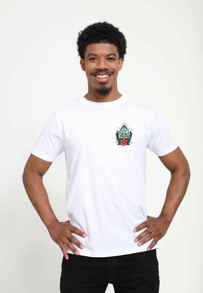 Hvid bomulds t-shirt med et farverigt grafisk design på venstre bryst, der viser en karakter blandt blomsterdetaljer. Korte ærmer, klassisk pasform.