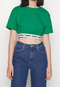Vihreä lyhythihainen t-paita, joka on valmistettu puuvillasta, pyöreällä kaula-aukolla ja mustalla vyötärönauhalla, jossa lukee "TOMMY JEANS". Yhdistettynä sinisiin farkkuihin.