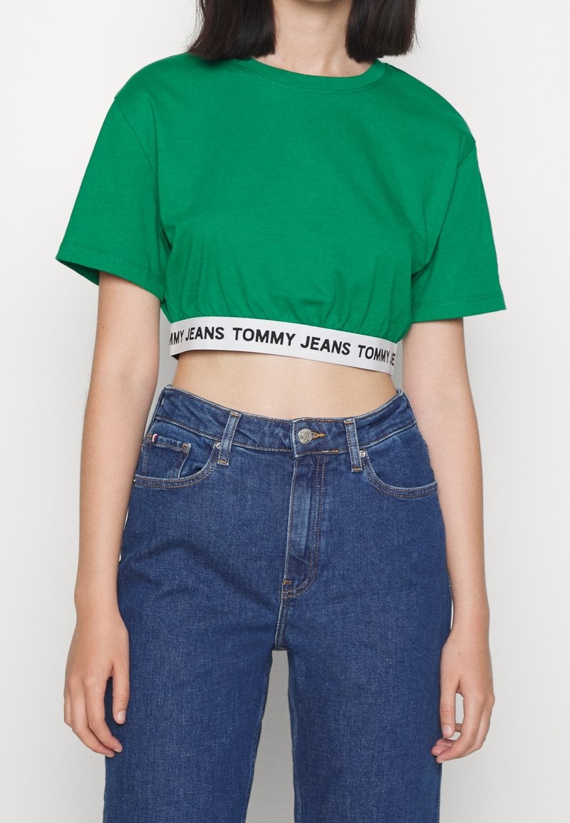 Vihreä lyhythihainen t-paita, joka on valmistettu puuvillasta, pyöreällä kaula-aukolla ja mustalla vyötärönauhalla, jossa lukee "TOMMY JEANS". Yhdistettynä sinisiin farkkuihin.