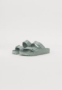 Des sandales en plastique vert avec deux brides réglables, une semelle texturée et une semelle plate. Le design est minimaliste et fonctionnel.