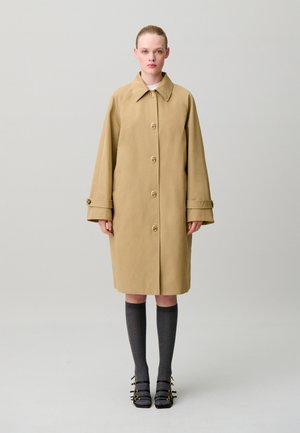 Mujer de pie vistiendo un abrigo beige hasta la rodilla con grandes botones, calcetines grises hasta la rodilla y zapatos negros con hebilla sobre un fondo claro y liso.