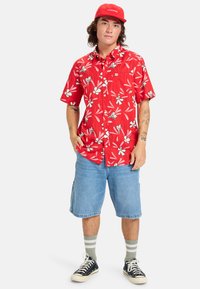 Camisa roja de manga corta con un estampado floral blanco, combinada con pantalones cortos de mezclilla azul claro. La persona lleva una gorra roja, calcetas rayadas grises y zapatillas negras.