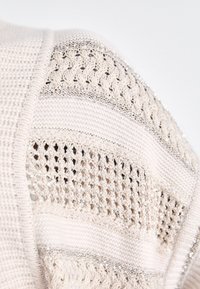 Strickpullover mit beiger Textur, der abwechselnde Muster aufweist: offenes Gewebe, durchgehende Streifen und metallische Fäden, die über den Stoff verlaufen.