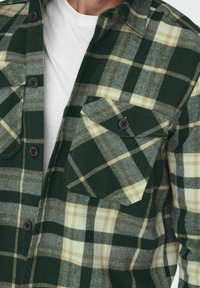 Camisa de franela a cuadros en verde y crema con cierres de botón, dos bolsillos en el pecho y un patrón texturizado, que presenta un cuello descentralizado y mangas largas.