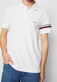 Vit polo t-shirt med en klassisk krage och tre knappar. Har en randig accent i röd och marinblå på ärmarna. Mjuk bomullstyg.