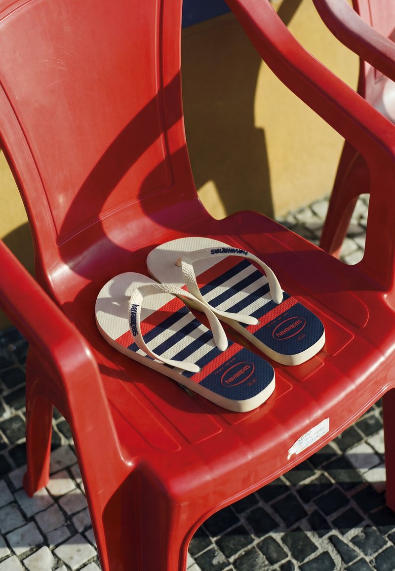Paire de tongs blanches, rouges et bleues rayées sur une chaise en plastique rouge sur une surface extérieure carrelée au soleil.