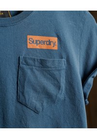 Modrá krátkosrstá tričko vyrobené z bavlny, s kapsou na prsou a oranžovým obdélníkovým logem "Superdry" na hrudi.
