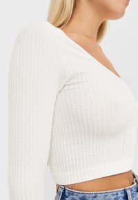 Biały długi rękaw crop top z fakturowanej, prążkowanej tkaniny i dekoltem w kształcie litery V. Dopasowany krój uzupełniony wysokim stanem dżinsów.
