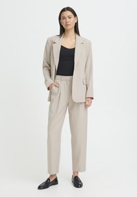 Blazer beige sur mesure avec un seul bouton, associé à un pantalon beige taille haute. Débardeur noir en dessous, mocassins noirs complètent le look.