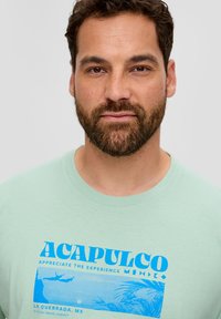 Jasnozielona bawełniana koszulka z niebieskim nadrukiem z napisem "ACAPULCO" oraz tropikalną sceną i detalami związanymi z biurem podróży.