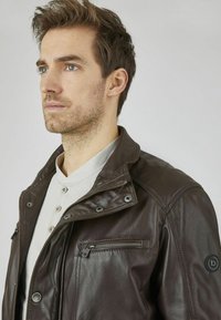 Braune Lederjacke mit einem Reißverschlusstasche und Druckknöpfen, ausgestattet mit einem klassischen Kragen und strukturiertem Finish. Trägt man über einem hellgrauen Hemd.