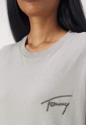 Kvinde iført en lysegrå t-shirt med rund hals og mørk broderi med ordet "Tommy" på brystet.
