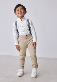 White button-up shirt, beige pants with suspenders, and white sneakers. Textured fabric, fitted design, with rolled cuffs and visible stitching.