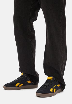 Czarne zamszowe trampki z żółtymi akcentami, wyposażone w płaską gumową podeszwę, sznurowadła i logo na języku, zestawione z czarnymi, luźnymi dżinsami.