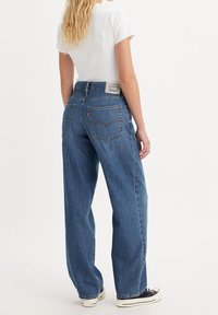 Žena s dlouhými blond vlasy oblečená v bílém tričku, volných modrých džínách a černo-bílých teniskách, stojící proti jednoduchému pozadí.