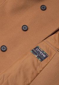 Cappotto di lana marrone con design doppiopetto. Bottoni neri, fodera interna liscia e un'etichetta con dettagli verdi e neri.