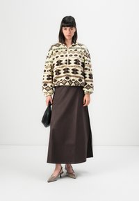 Vrouw in een geblokte fleece pullover, lange donkerbruine rok, met een zwarte handtas en beige puntige hakken, staand op een witte achtergrond.