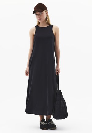 Giovane donna con un abito lungo nero senza maniche, cappello marrone scuro, sneakers nere e una borsa trapuntata nera in mano.