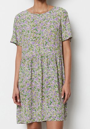 Vrouw die een losse jurk met korte mouwen draagt, met een groen, paars en wit bloemenpatroon, staand voor een effen achtergrond.