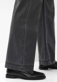 Zwarte loafers met een glanzende afwerking, gecombineerd met grijze jeans met wijd uitgesneden pijpen, die een gladde textuur en een gestikte zoomafwerking hebben.