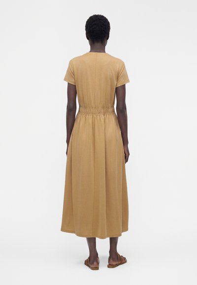 Femme debout, dos visible, portant une robe midi à manches courtes couleur chameau avec une taille élastique et des sandales plates marron.
