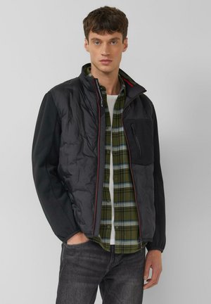 Jonge man die een zwarte zip-up jas draagt over een groen geruit overhemd en een wit ondershirt, met donkere spijkerbroek, poseert tegen een effen grijze achtergrond.