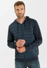 Sudadera con cremallera de color azul oscuro, con panel frontal acolchado, mangas grises, ribete a rayas rojas y blancas en la capucha y un pequeño parche de logo.