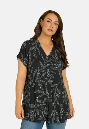 Vrouw met lang bruin haar, gekleed in een zwart kort mouw V-hals top met witte bloemmotieven en een blauwe spijkerbroek.
