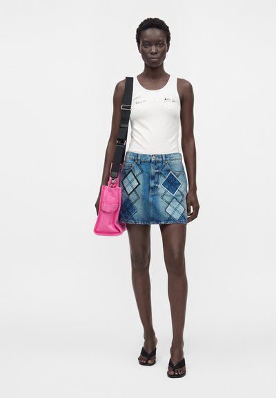 Femme portant un haut blanc sans manches, une jupe en jean bleue avec des motifs en diamant, des sandales noires, et portant un sac à bandoulière rose vif.